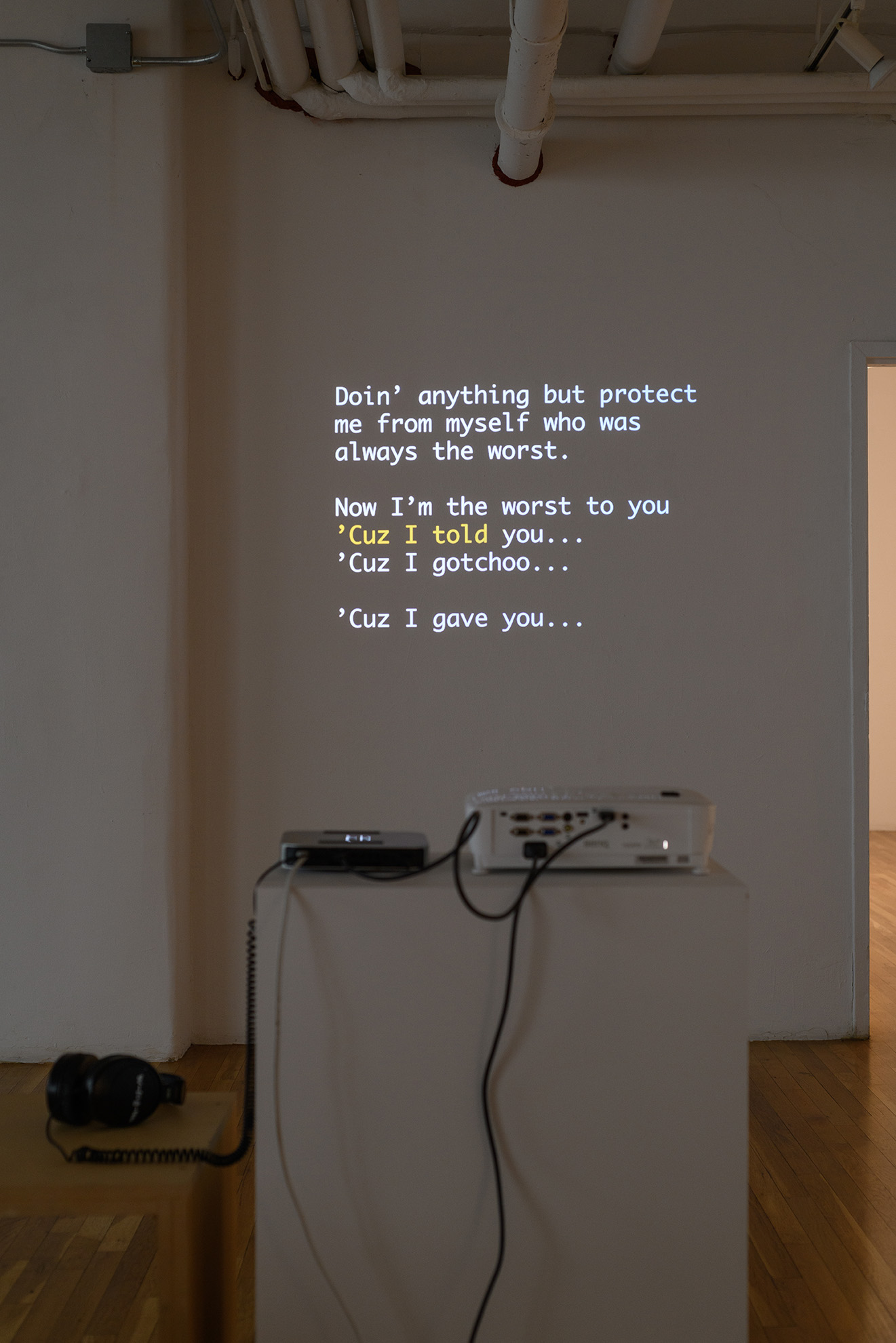 [Another view of the same projection at a different point in the video showing both the projected image and the projector machine, media player, and cabling–all sitting on a white plinth. The projected image shows song lyrics in white text with a portion highlighted in yellow, as is true of karaoke videos. It reads, quote: “Now I’m the worst to you / ‘Cuz I told you… / ‘Cuz I gotchoo… / … / ‘Cuz I gave you… ]